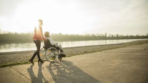 Westend61/Getty Images  A woman pushing another in a wheelchair