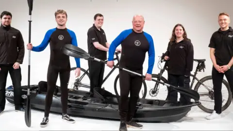 Two men in black an blue wet suits holding a paddle, There are three men and one woman dressed in all black smiling looking at the two gentleman. 