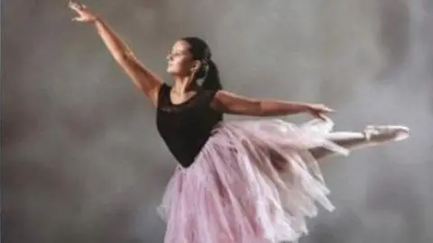 Family supplied A young female dancer in profile. She is standing on the top of her toes on her right leg with her left leg out behind her. Her right arm is extended in front of her and her right behind her. She is wearing a calf-length light-pink coloured tutu skirt and black vest top. She has long dark hair pulled back from her face in a ponytail.