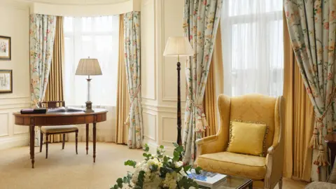 Pro Auction A room inside The Savoy with floral curtains, a wooden desk and yellow armchair.