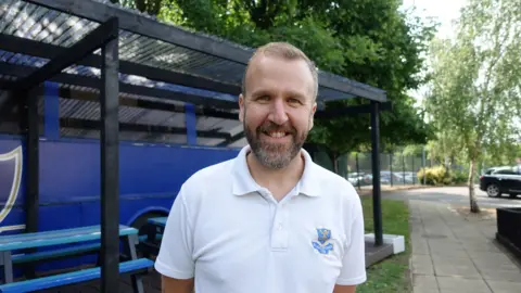Sam Brunt in front of the book bus, wearing a white polo shirt with the school crest on it.