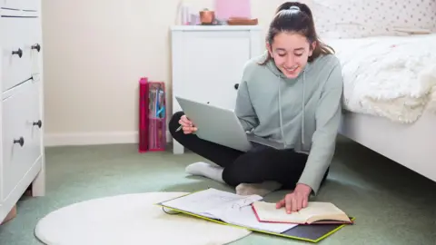 Getty Images A girl wearing a light green jumper with brown hair tied up in a ponytail, she is sat on her bedroom floor with her laptop on her legs and her folder and notebook on the floor, she is smiling and is reading her notes. 
