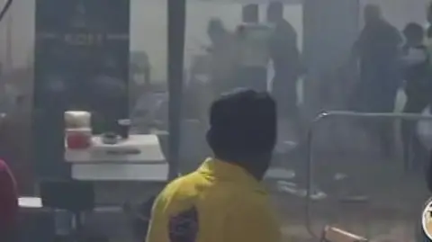 The back of a man wearing a yellow shirt looking at a smoke filled area. There are people in the background.