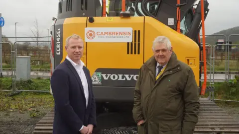 Herefordshire Council A man in a navy blue suit and white shirt stands beside a man in a dark green coat. In the background is a yellow Volvo digger and metal fences. 