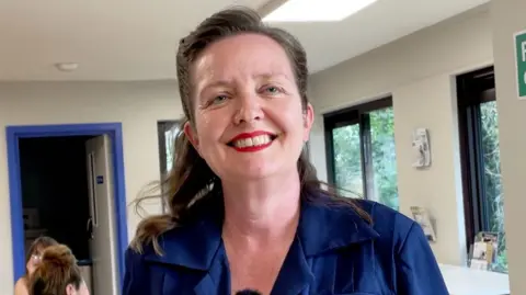 Kit Taylor/BBC A woman with brown hair in a blue dress with white buttons. She is smiling at the camera.