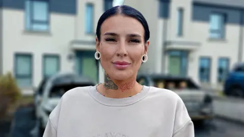 Elise Conroy is a young woman with slicked back dark hair and stretchers in her ears. She is smiling at the camera. She is wearing a cream jumper.