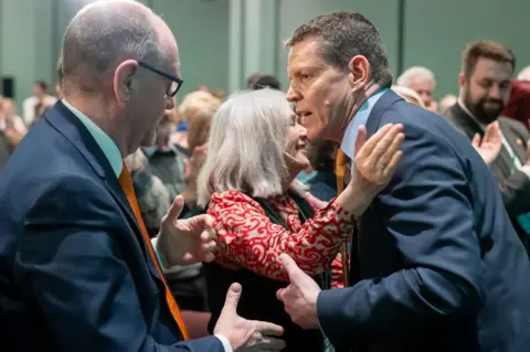 Matthew Horwood A man, Rhun ap Iorwerth, is embraced by a woman with grey hair while people around them applaud.