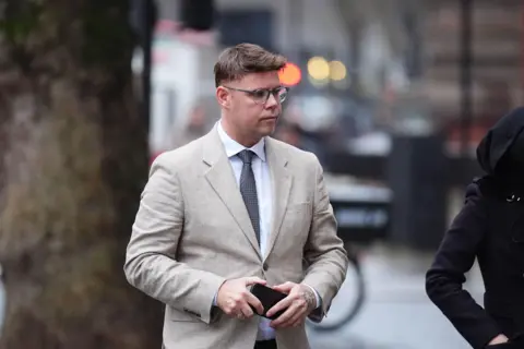 PA Media Neil Debenham wearing a greige suit and tie and holding his phone in his hands