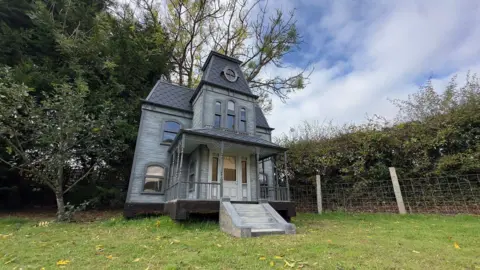 A large 12ft grey wooden Halloween house, with sloped roofs and small arched windows. There is a grey staircase leading up to a wooden porch and white front door. There is a circular window on the roof at the front