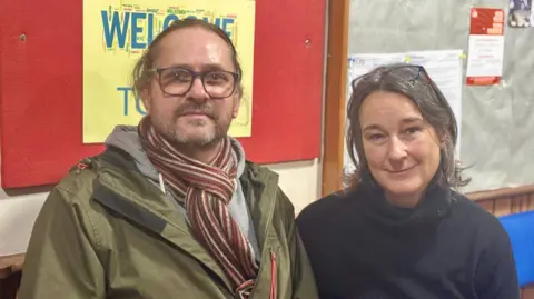 A man with long hair and a khaki jacket wears a stripey red and cream scarf. He is sitting next to a woman in a navy roll necked jumper - she has dark hair and glasses on her head