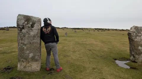 Dean Cooper Dean Cooper on a walk in Cornwall which helps him deal with burnout and stress. He is seen in a black jumper and grey jeans leaning against a rock while looking out to a view of a green field.