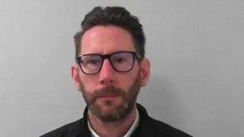 A custody picture of a man with short dark hair, a beard, and glasses, wearing a white collared shirt under a dark sweater and a black quilted jacket, standing against a plain light grey wall.