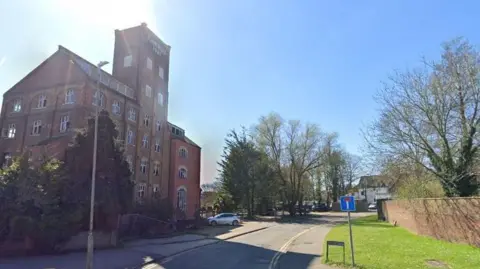 A Google Streetview image showing East Hill in Ashford which has a large old mill building on the left hand side and other buildings and trees seen further up the street. There is a small patch of grass on the right of the image, a small street sign and a red brick wall is also seen.