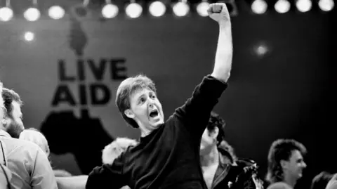 A black and white photo of former Beatle Paul McCartney saluting the audience at Live Aid. He is wearing a black top and the Live Aid logo is in the background.