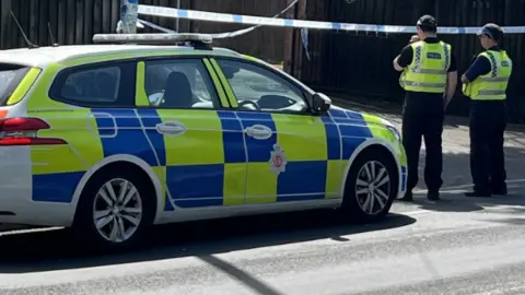 A police car and two police officers are stood outside crime tape which is in front of a large gate, in a residential area. 