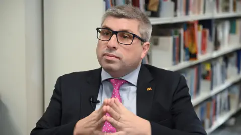 Mark Arnull in a navy blazer, blue shirt and pink tie sits in front of a book shelf looking off to the left of camera.