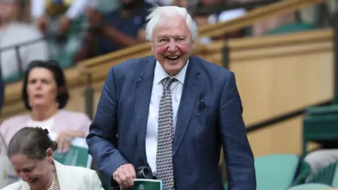 Getty Images Sir David Attenborough na Wimbledonie. Uśmiecha się, siadając na swoim miejscu, ma na sobie ciemnoniebiesko-szarą marynarkę i czarno-biały krawat ze wzorem