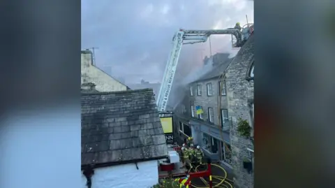 Eugene Kaufman Firefighters on the street in front of the building that caught fire in Kirkby Lonsdale. They are working with a colleague who is on an aerial ladder to bring the flames under control.
