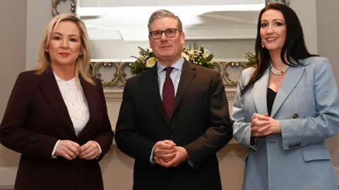 PA Media Three people in formal wear posing for a photograph inside in front of a mirror. The man in the middle has grey hair is wearing glasses. The woman on the left has blonde hair and the woman on the right has brown hair.
