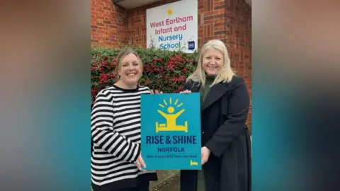 Sara Kenny Sara Kenny wears a dark coat and holds a large small board with Jade Kenny. The board is aqua blue with dark blue writing, which reads "RISE AND SHINE NORFOLK". The board also has a drawing in yellow of a person sitting up in bed with their arms outstretched. The pair are stood outside a building of West Earlham Infant and Nursery School and a sign on the brick building behind them indicates this. There is a hedge with red berries behind the women. 