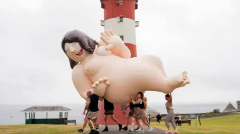 The Box A naked inflatable character - based on the Beryl Cook painting Nude on a Leopard Skin - is being carried across Plymouth Hoe's waterfront, in front of the red and white lighthouse Smeaton's Tower.