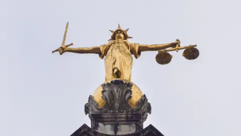 A gold statue on top of the Old Bailey from below. It is of a woman in a dress with a spikey hat and outstretched arms. In one hand she is holding some scales and the other a sword. The sky above is grey.