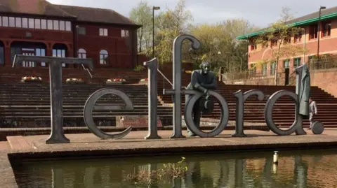 A metal sign spelling out the letters to T-E-L-F-O-R-D stands on a paved area next to a small pond. Behind it are stone steps leading to brick buildings. Over the letter "o" of the sign, leans a sculpture of a man.