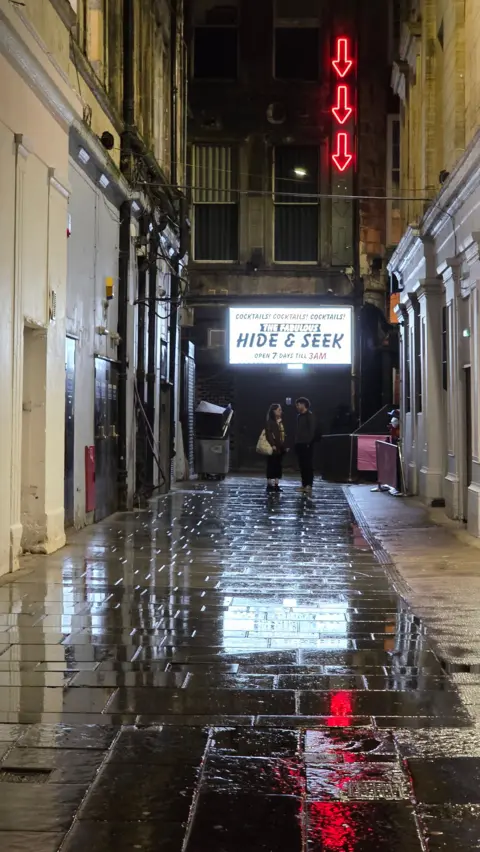 Sarah Robertson A city street, illuminated by a large white sign reading HIDE & SEEK, while another sign is lit with downward red arrows. A man and a woman are standing underneath them talking. 