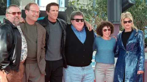 Alamy Cheers co-stars standing in a row with with their arms around each other in 1999. Left-right: John Ratzenberger, Kelsey Grammer, Ted Danson, George Wendt, Rhea Perlman and Kirstie Alley.