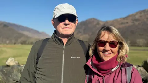 Mike Butler standing next to his wife. He's wearing a blue cap and sunglasses and green jacket. His wife has blonde hair and is smiling into the camera.