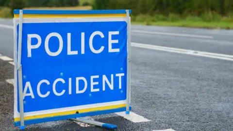 A blue and white sign, positioned on a country road, reads "Police Accident".