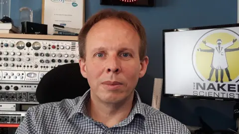 Chris Smith, a man with light brown hair, wearing a blue and white checked shirt. He is sitting in front of radio recording equipment. A screen over his left shoulder is showing The Naked Scientists logo of a man in a white coat with his arms outstretched.
