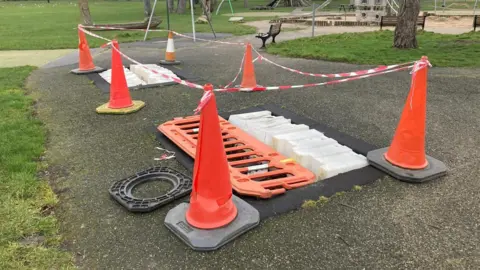 A children's playpark which has a section cordoned off with red traffic cones, red plastic gating and red and white taping. The gating is placed over two holes in the ground where trampolines once were. 