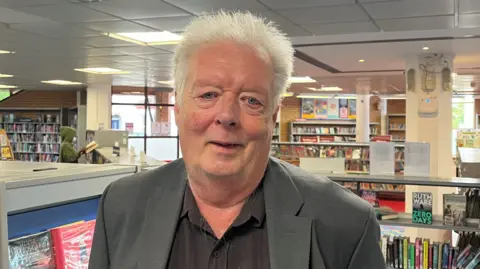 Barnet Council leader Barry Rawlings looking at the camera. He is standing inside Chipping Barnet Library with shelves of books behind him