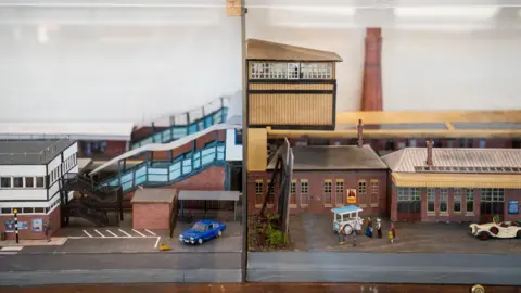 Great Western Railway A model railway showing Gloucester station in two different time periods. The left side of the display represents a more modern era, with a more modern-looking car, while the right side shows an earlier, more industrial version of the station with an older model car.