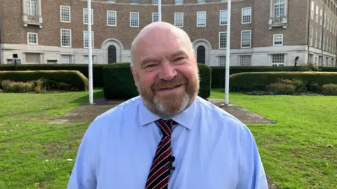 A man with a beard is wearing a shirt and tie, and is standing in front of a council building