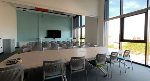 An empty classroom with a large desk in the centre which is surrounded by rows of grey and metal chairs. There is a large video screen at the end of the room and on the right side of the building is a floor to ceiling glass window.