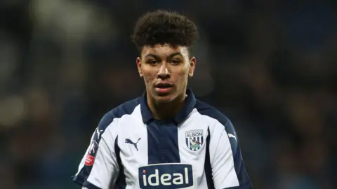 Getty Images A young Morgan Rogers is wearing a West Bromwich Albion kit. He has short hair which is shaved on the side. Behind him is blurred but there is a crowd. He is looking ahead and not at the camera. 