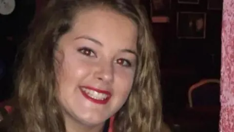 Emily smiles at the camera. She has long wavy brown hair and red lipstick on.