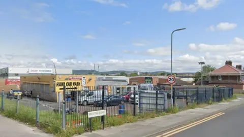 Hutton Road. There is a yellow sign reading Hand Car Wash behind a fence on the pavement.  A garage changing tyres is further away with a number of cars parked alongside it. There is 20mph speed restriction sign. 