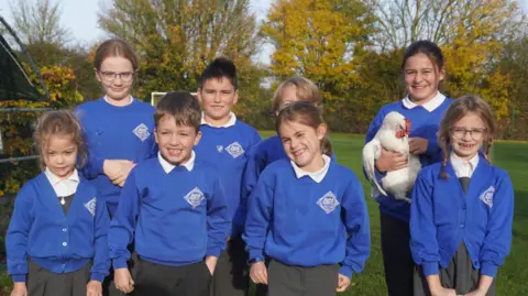 St Edmundsbury and Ipswich Diocesan Multi-Academy Trust A small group of pupils stand in a group to pose for a photo. Taller and older students stand in a line behind another line of smaller younger students. One of the older students is holding a white hen. They are all wearing blue school jumpers with white shirts underneath.