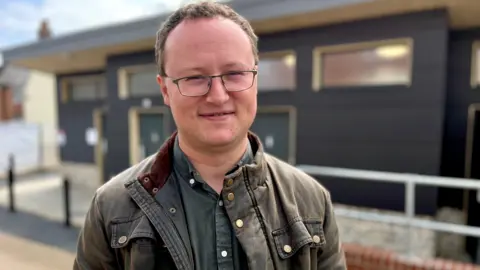 A man with glasses wearing a wax jacket and green shirt smiles while looking towards the camera.