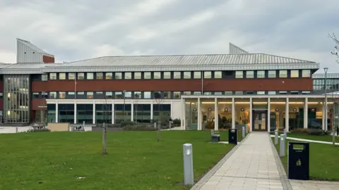 County Hall in Morpeth. It is a large office block with lots of windows, red brick walls, a glass atrium at the front and a silver slanted roof.