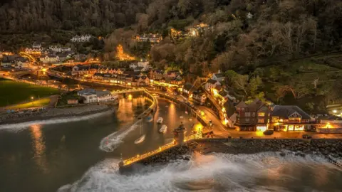Photo provided by the South West Coast Path Association, Photographer: Shaun Davey An aerial shot over a seaside village, all lit up by the warm glow of orange street lights. Waves are crashing against a sea wall. Properties are lined along the harbourside. Trees are behind the properties. It is dusk.