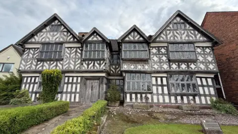 A black and white, three-storey Elizabethan building. There is a flagstone path leading up to a wooden door. There is a low hedge either side of the path. In front of the building is a small lawned area. There is a plaque in the middle of this.