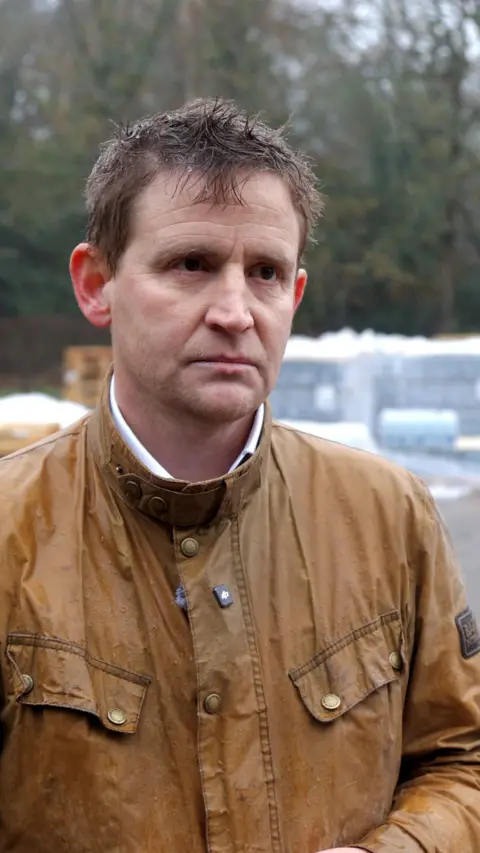 A man in a brown coat. There are crates of bottled water behind him.