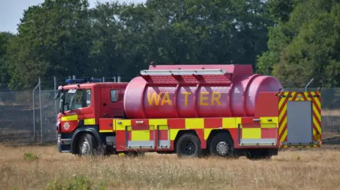 Richard Martin A red fire brigade water carrier is in a dry grass field. Green trees edge the field and the water truck has stopped outside a metal fence.