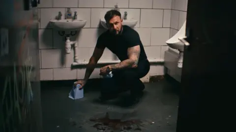 Hustle and Cuss/Jack Grewar Luke is crouching in a washroom in a still image taken from the Hustle and Cuss film