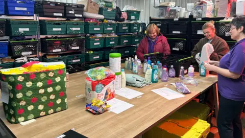 A photo showing three women stood around a table packing items into bags. In the background are trays of items stacked up