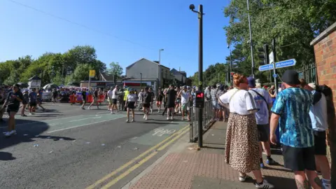 Emma Gibbs Oasis fans queue by the side of a wall that marks the boundary of Heaton Park, while others cross a road from the Metrolink stop to the park in the background